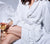 Woman sitting on bed dressed with Essential White Percale sheets, holding a White Wine Glass and wearing the Essential White Classic Bathrobe.