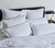Dark wood bed dressed in white bedding, blankets pulled down and six Down Pillows with pillowcases.  Side table with lamp.