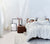 Woman lying in bed with linen sheets and duvet pulled up.  Framed art and side table with decorative items and flowing white curtains.