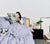 Woman sitting in bed dressed with Slate Blue Linen Sheets and Duvet reading a newspaper and having a snack. Side table with other decorative items.