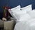 White bed with 6 pillows in a dark blue painted room. Silver bedside table with decorative objects and flowers on it.