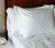 Wooden bed dressed in Essential White Percale Sheets with Down Alternative Pillows inside a Pillow Case and Pillow Sham. Side table with candle on it next to bed.