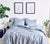 Slate Blue Percale bed with four pillows. Plants, lamp and other decorative objects around it. Two pictures hanging on the wall. Plants and decorative objects around it.