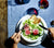 Person eating a salad from our white porcelain Dinner Bowl sitting atop of Dinner Plates.  Red Wine Glass and Classic Stainless Flatware are also featured on a blue table cloth.